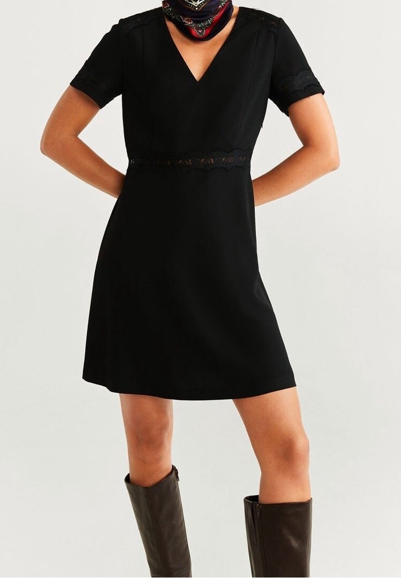 Femme portant une robe noire à manches courtes avec un col en V orné de dentelle et des bottes marron foncé montant jusqu'aux genoux, debout les mains sur les hanches.