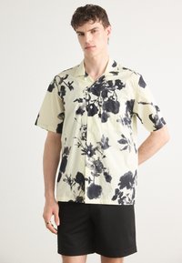 Camicia a maniche corte, con bottoni, di colore giallo chiaro con stampe floreali nere; tessuto morbido, vestibilità comoda, con taschino sul petto.