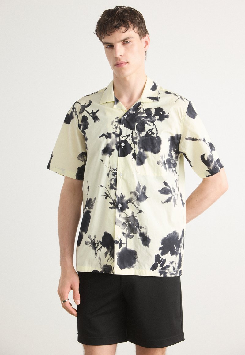 Camicia a maniche corte, con bottoni, di colore giallo chiaro con stampe floreali nere; tessuto morbido, vestibilità comoda, con taschino sul petto.