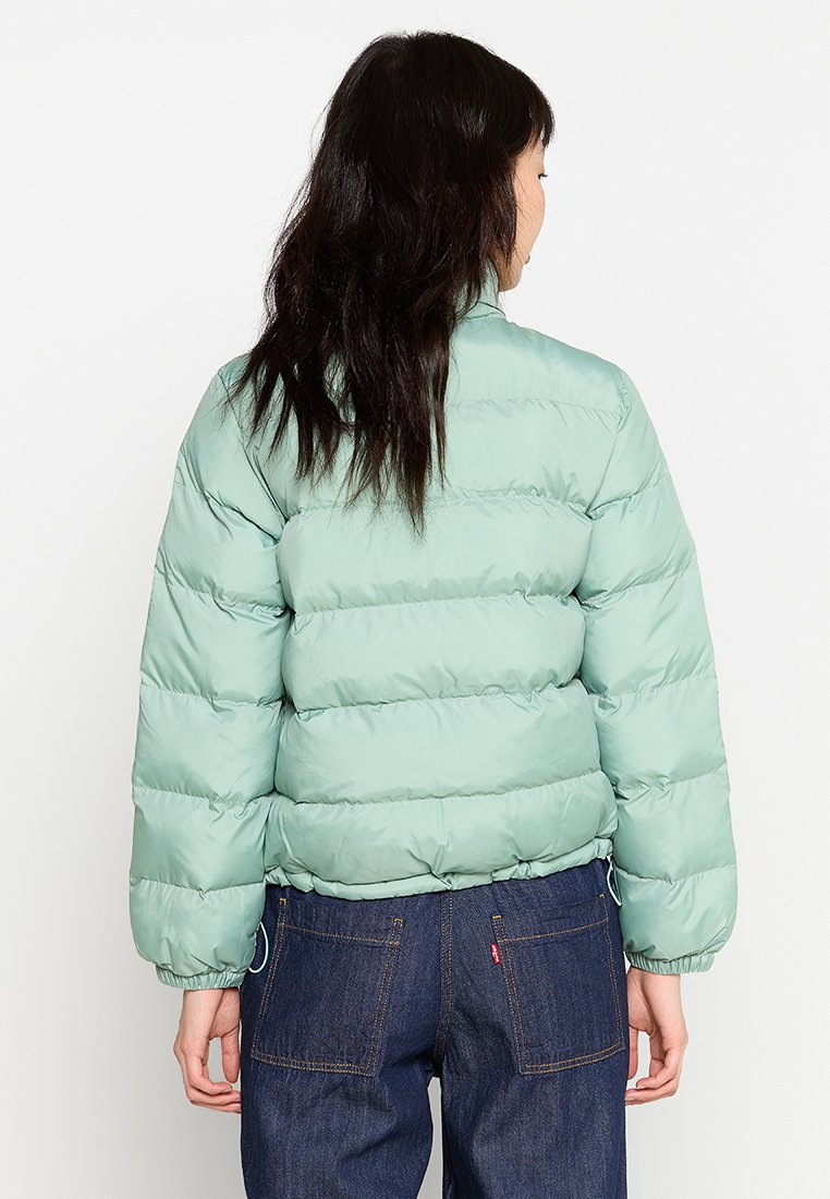 Chaqueta de plumas verde claro con costuras horizontales, puños elásticos, cuello elevado y dobladillo metido, llevada sobre unos vaqueros oscuros.