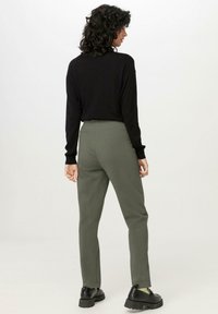 Schwarzes langärmeliges Oberteil, kombiniert mit grünen, schmal zulaufenden Hosen, die einen elastischen Bund und schwarze klobige Schuhe aufweisen, von hinten betrachtet.