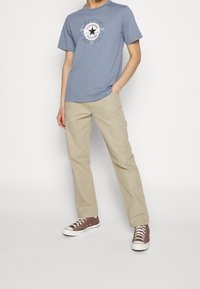 Light blue cotton t-shirt with a round neck and a graphic logo design, paired with beige cotton trousers and brown canvas sneakers.