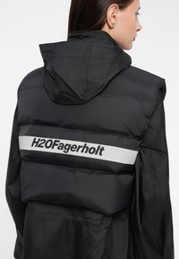 Gevoerde zwarte jas met een capuchon en een zilveren streep met de tekst "H2OFagerholt", die een elegante, gestructureerde oppervlakte toont.