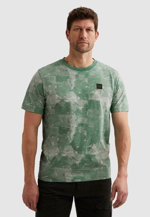 Mann trägt ein kurzärmeliges grünes gemustertes T-Shirt und schwarze Hose, steht vor einem einfarbigen hellen Hintergrund.