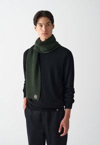 Fular verde cu textură înfășurat în jurul gâtului, realizat dintr-un material moale; purtat peste un pulover negru; are un accent cu un mic logo aproape de capăt.