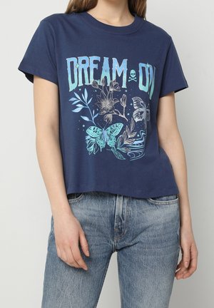 Femme portant un t-shirt bleu marine avec le texte "DREAM ON", des papillons bleus, des fleurs et des motifs étoilés, associé à un jean bleu clair.
