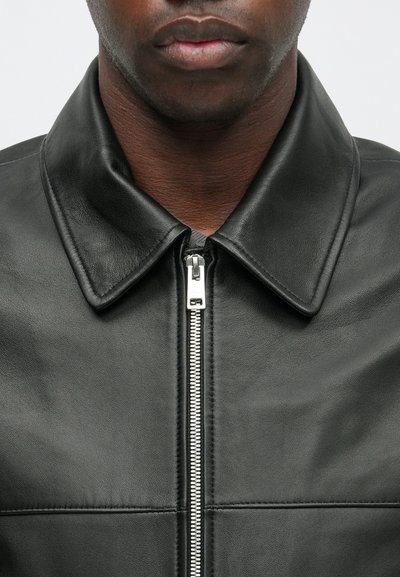 Giacca in pelle nera con colletto, dotata di una zip argentata e dettagli cuciti nella parte anteriore. Texture liscia e design aderente.