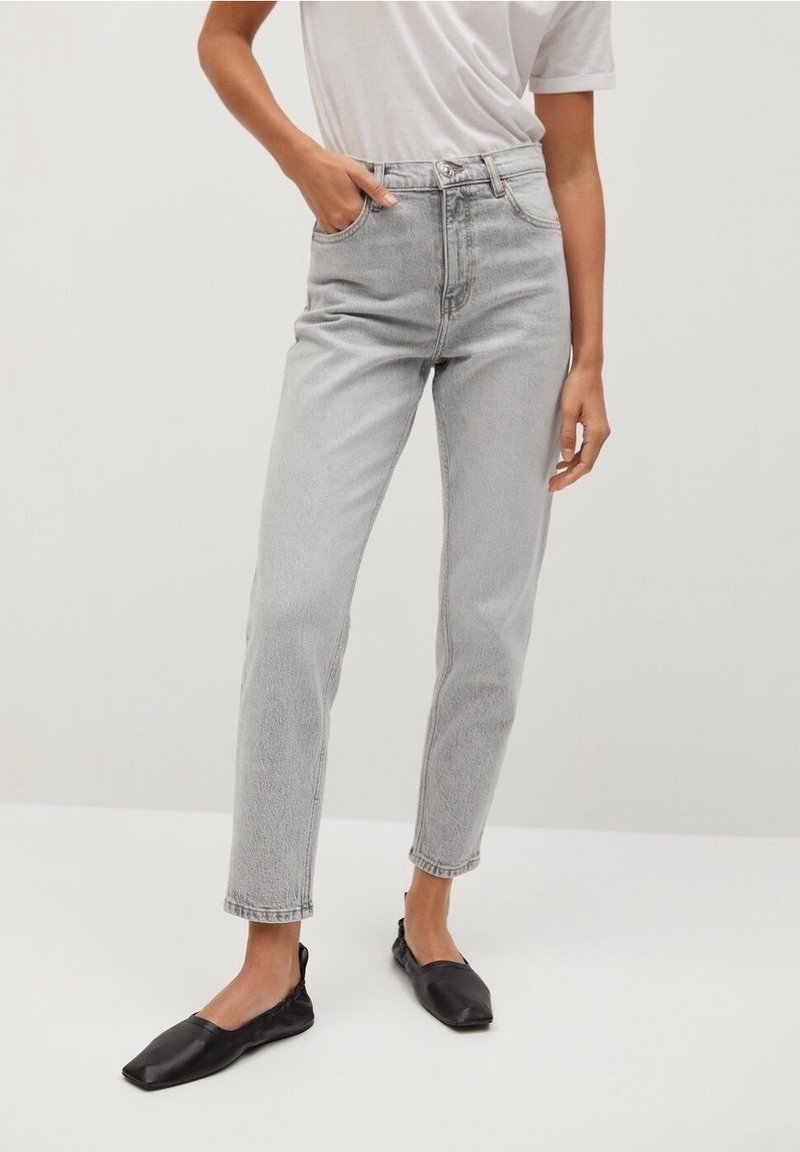 Personne portant un jean droit taille haute gris clair avec un T-shirt blanc rentrée et des chaussures plates noires à bout carré, debout avec une main dans la poche.