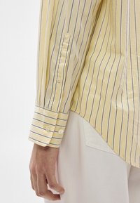 Dzeltena svītraina krekls ar šaurām zilām līnijām, ar pogām aiztaisāmām aprocēm, izsmalcinātu zīmolu uz piedurknes un nedaudz izliekta apakšmala.