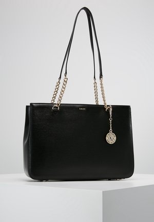 Schwarze DKNY Ledertasche mit goldenen Kettenriemen und rundem goldenen Logoanhänger, präsentiert auf einem weißen Podest vor grauem Hintergrund.
