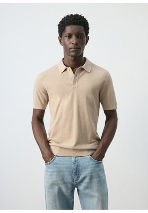 Hombre con cabello corto y rizado, vestido con camiseta tipo polo beige de manga corta y jeans azul claro, de pie con las manos en los bolsillos sobre un fondo liso.