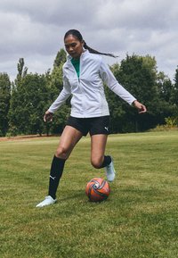 Hvit atletisk jakke, grønn topp, svarte shorts og knelangne svarte sokker. Sparkende en flerfarget fotball på en gresskledd bane.