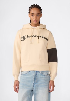 Person trägt einen beigen Champion-Hoodie mit schwarzem Logo auf der Brust und einem schwarzen Fleck an einem Ärmel, kombiniert mit hellblauen Jeans.