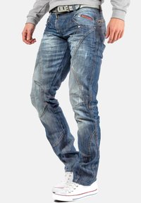 Jean en denim bleu avec une finition délavée, des détails de couture complexes, une coupe cintrée et un design comportant de nombreuses poches. Associé à des baskets blanches.