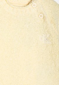 Pull en polaire jaune avec une texture douce, un col côtelé et trois boutons à l'épaule ; comprend un logo brodé blanc.