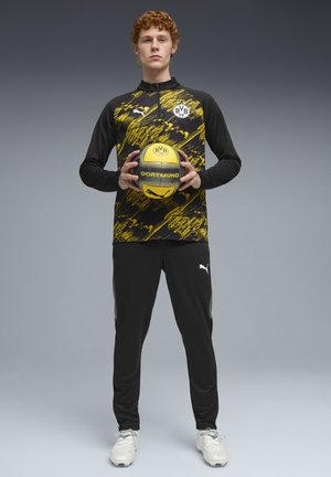 Joven con chaqueta negra y amarilla del Borussia Dortmund y pantalones negros sostiene un balón de fútbol amarillo y negro con el texto "Dortmund".