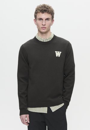 Dark green knitted sweater with a round neckline, ribbed cuffs, and hem, featuring a white "W" logo on the chest, layered over a green striped shirt.