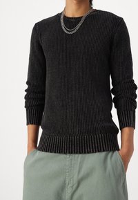 Personne portant un pull côtelé foncé, un collier chaîne en argent et un pantalon vert clair, les mains dans les poches, devant un fond uni.