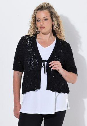 Kvinde med krøllet blondt hår iført en sort kortærmet strikket cardigan over en hvid skjorte og sorte bukser, stående mod en ensfarvet baggrund.