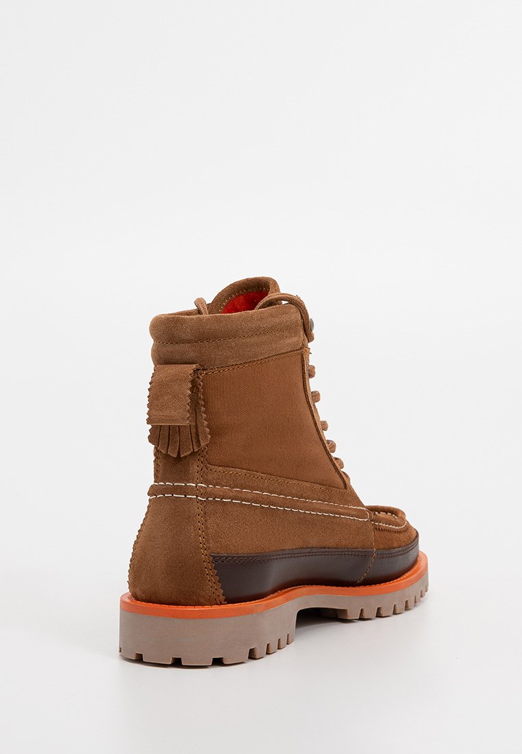 Braune Wildlederstiefel mit hohem Schaft, weißer Naht, bordeauxroter Innenauskleidung und einer robusten orange-grauen Sohle. Mit einem Quastenakzent.