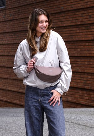 Tânără zâmbitoare, purtând o jachetă gri deschis, blugi albaștri și o geantă maro purtată în diagonală, stând în fața unui zid de cărămidă.