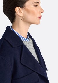 Cappotto di lana blu navy con grandi rever, indossato sopra un maglione grigio e una camicia a righe blu. Orecchini a cerchio semplici completano il look.