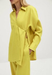 Lime vihreä pellavapaita, jossa on kauluksellinen pääntie, pitkät hihat, sivu solmudiili ja epäsymmetrinen helma. Sileä pinta ja hengittävä materiaali.