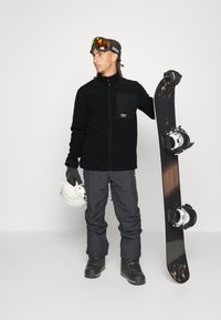Sort fleecejakke med lynlås lomme, grå snebukser, sorte handsker, hvid hjelm og snowboard med bindinger, båret af en person.