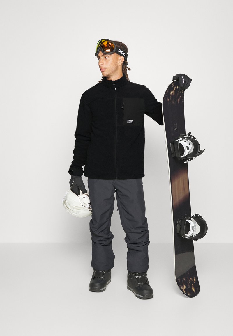 Chaqueta de forro polar negra con bolsillo con cremallera, pantalones de nieve grises, guantes negros, casco blanco y tabla de snowboard con fijaciones, llevados por una persona.
