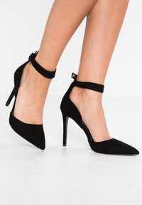 Černé semišové lodičky na vysokém podpatku s špičatou špičkou, řemínkem kolem kotníku a otevřenými stranami, s lesklým podpatkem a hladkým matným povrchem.