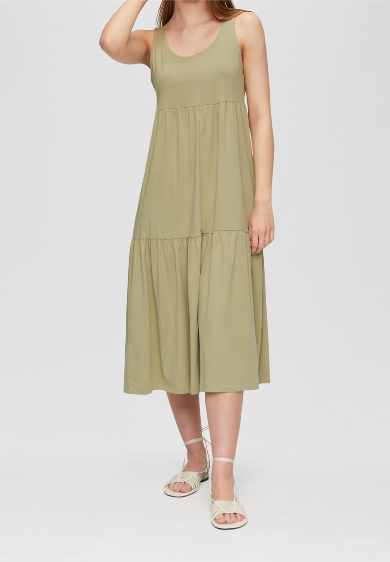 Femme portant une robe midi sans manches, ample, vert olive clair, et des sandales blanches à brides, debout devant un fond uni et clair.