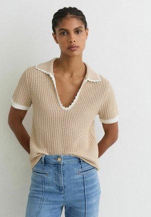 Kobieta z zaplecionymi włosami ubrana w beżowy, dziergany top z krótkim rękawem z białą lamówką oraz jasnoniebieskie, wysokie spodnie dżinsowe, stojąca przy białej ścianie.