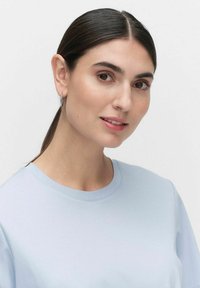 Vaaleansininen, lyhythihainen paita, jossa on pyöreä pääntie. Mallilla on yllään vähäiset hopeiset korvaketjut ja suorat tummat hiukset kiinni.