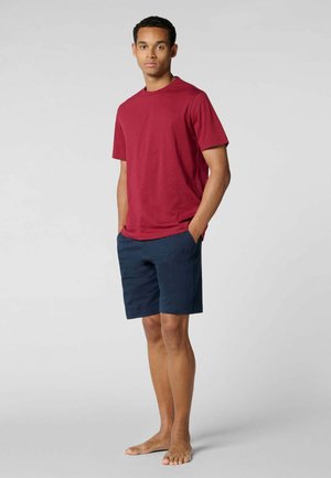 Giovane uomo rilassato in piedi a piedi nudi che indossa una maglietta rossa a maniche corte e pantaloncini blu navy su uno sfondo chiaro e uniforme.