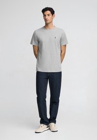 Grijze katoenen t-shirt met korte mouwen, ronde hals en een klein zwart logo geborduurd op de linkerborst; gecombineerd met donkerblauwe spijkerbroek en beige schoenen.