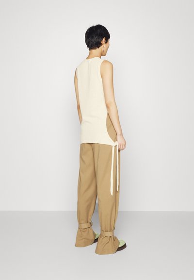 Beige ujjatlan felső oldalsó sliccekkel és megkötőkkel, bézs laza nadrággal, összegyűrt bokákkal és világoszöld szandálokkal párosítva.