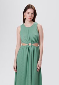 Vestido verde menta con tejido texturizado, diseño sin mangas y corte circular en la cintura. La falda fluida se extiende hasta el suelo.