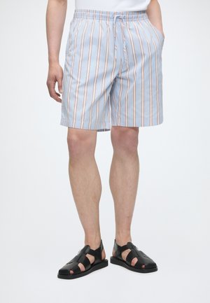 Άνδρας που φοράει ανοιχτό μπλε και λευκές ριγέ βερμούδες με πορτοκαλί λεπτομέρειες, λευκό πουκάμισο και μαύρα δερμάτινα σανδάλια, στέκεται με τα χέρια στις τσέπες.