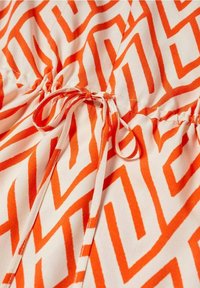 Robe avec un motif géométrique en zigzag orange sur un fond clair, avec une taille froncée et des rubans noués au centre pour un ajustement réglable.