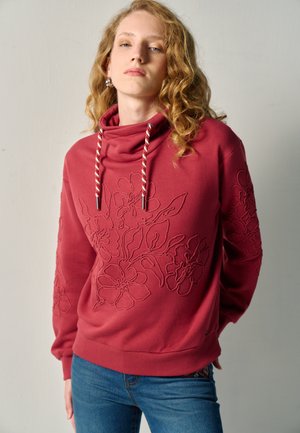 Giovane con capelli biondi ricci che indossa una felpa rossa con ricamo floreale e jeans blu, in piedi su uno sfondo neutro.