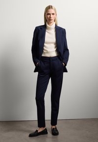 Blazer navy e pantaloni su misura abbinati a un dolcevita crema, caratterizzati da texture morbide e silhouette elegante. Mocassini neri completano il look.