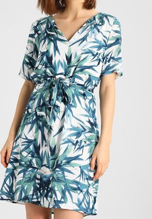 Donna con un abito a maniche corte, di lunghezza al ginocchio, con motivi di foglie verdi e blu e una cintura di tessuto legata in vita.