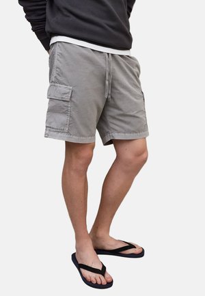 Person som har på seg grå cargo-shorts, svarte flip-flops og en mørk langermet genser, står foran en ensfarget hvit bakgrunn.