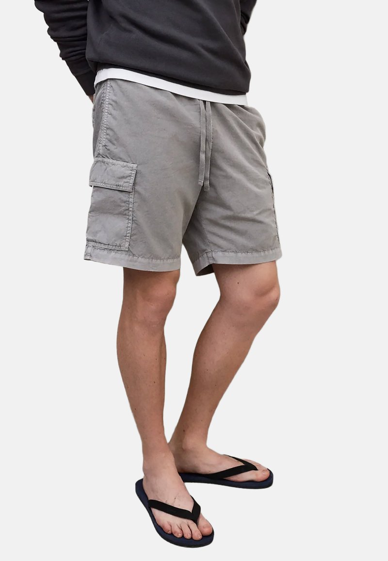 Persoon die grijze cargoshorts, zwarte slippers en een donker shirt met lange mouwen draagt, staand tegen een effen witte achtergrond.