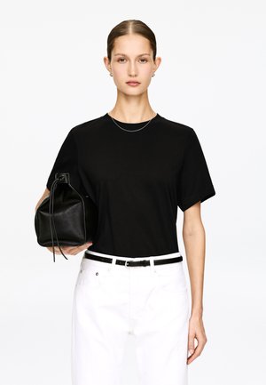 Femme aux cheveux lissés porte un t-shirt noir, un jean blanc avec ceinture noire, tenant un sac en cuir noir sur fond blanc.