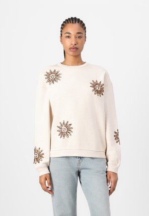 Light beige sweatshirt with long sleeves, featuring intricate golden sunburst embroidery with heart accents on shoulders and sleeves.