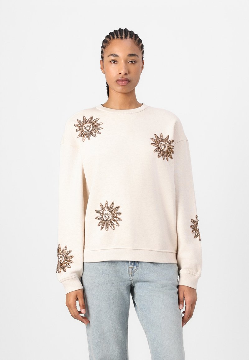 Light beige sweatshirt with long sleeves, featuring intricate golden sunburst embroidery with heart accents on shoulders and sleeves.