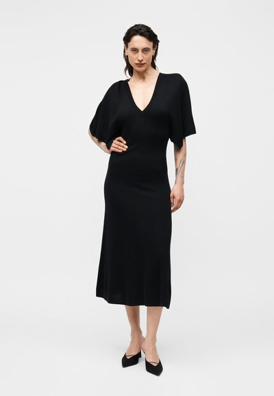 Γυναίκα με σκούρα μαλλιά που φοράει μαύρο φόρεμα μεσαίου μήκους με φαρδιά κοντά μανίκια και μυτερά μαύρα τακούνια, στέκεται μπροστά σε ένα απλό λευκό φόντο.