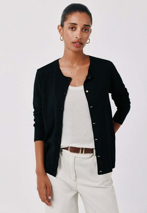Vrouw draagt een zwarte gebuttonede cardigan over een wit hemd, beige broek met een bruine riem en gouden oorringen tegen een lichte achtergrond.