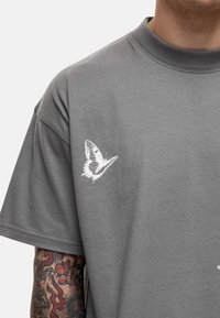 Grijze katoenen t-shirt met een witte vlinderborduursel op de linkerborst, met een losse pasvorm en korte mouwen.